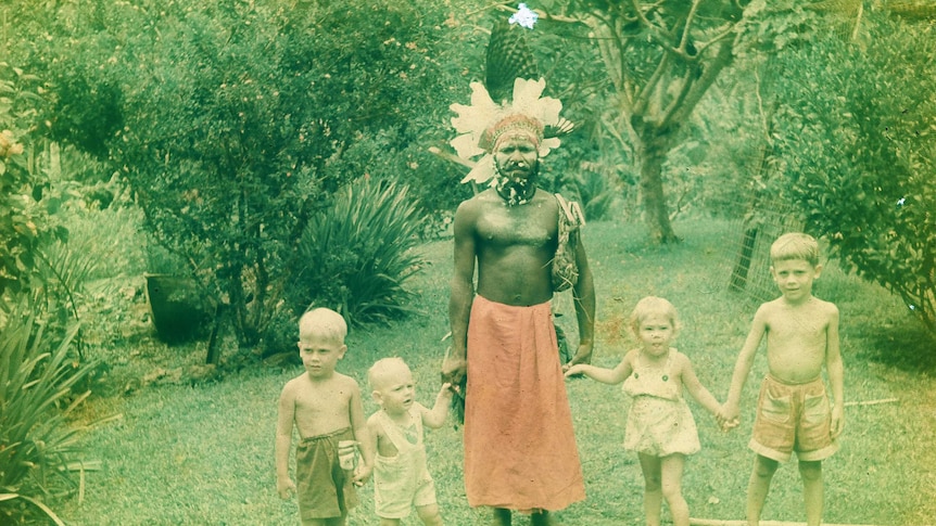 A faded photo shows four children holding hands in a row with a Papua New Guinean man in traditional dress in  a garden.