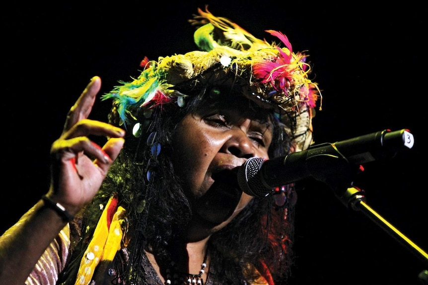 Ruby Hunter singing into a microphone wearing a colourful feathered head dress