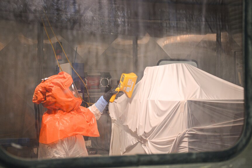 The worker is wearing a bright orange protective hood and holding a yellow electronic device