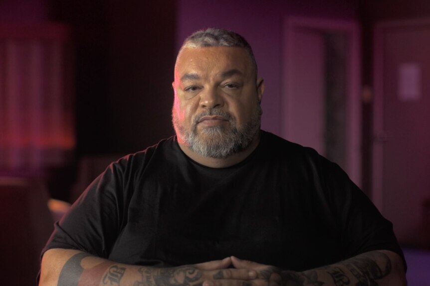 Head and shoulders image of an Indigenous man in a black t-shirt giving a tv documentary interview.