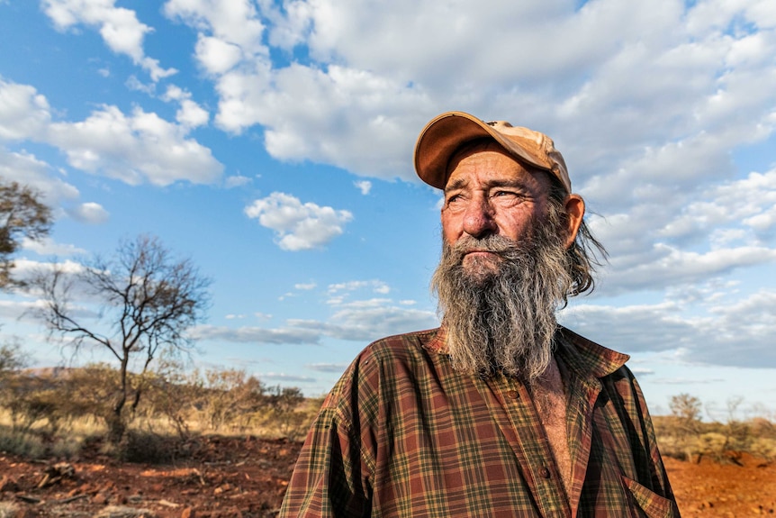 An elderly man with a beard and a baseball cap stands in the desert.