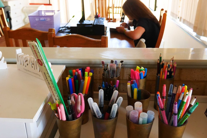 Cups filled with pencils and pens on a bench with a child sitting at a table doing homework in the background.