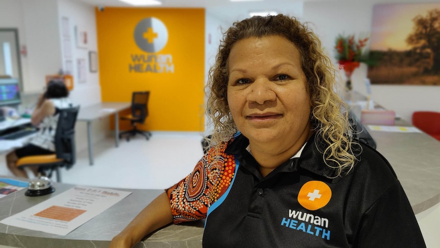 A woman with curly hair, wearing a t-shirt with Indigenous art on it, stands in a medical centre.