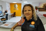 A woman with curly hair, wearing a t-shirt with Indigenous art on it, stands in a medical centre.
