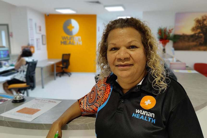 A woman with curly hair, wearing a tshirt with indigenous art print stands in a medical centre.
