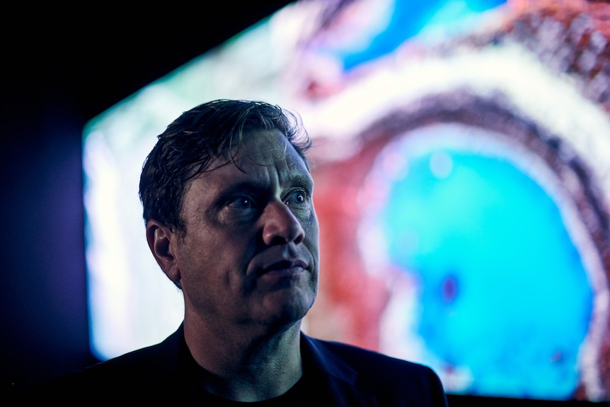 Profile shot of a 42-year-old white man with short, fair hair standing in a darkened room in front of a bright screen.