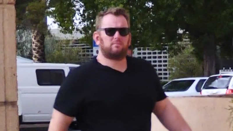 A bearded man with short hair wearing dark sunglasses and black shirt walks towards a courthouse.