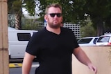 A man in a black t-shirt, jeans, and black sunglasses walking towards a court building.