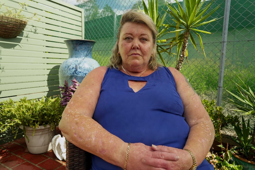 Woman with blonde hair wearing blue top sits outside in front of plants and a wire fence