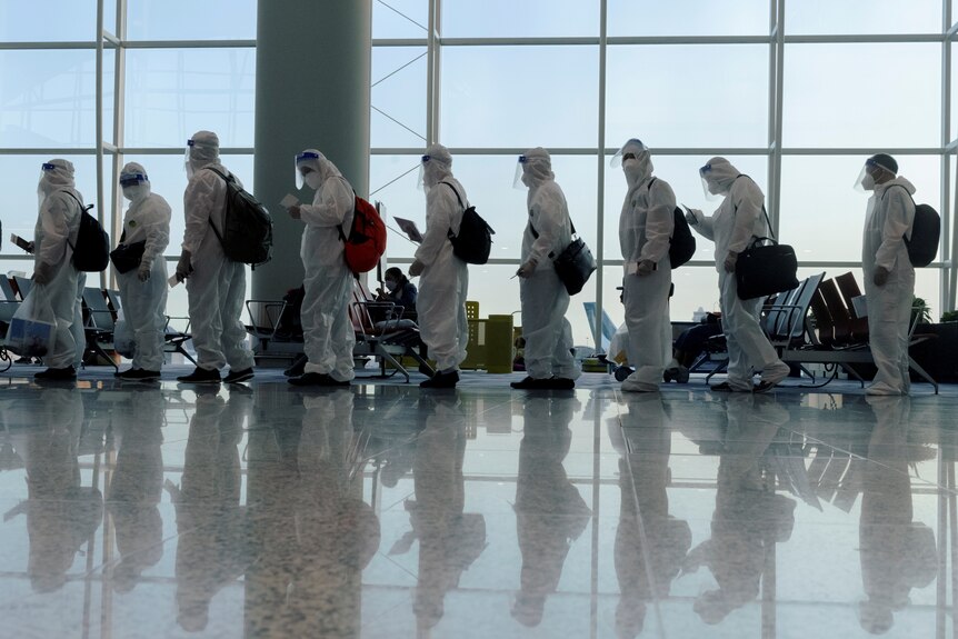A line of people all wearing full PPE, hazmat suits stand in an airport