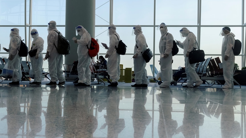 A number of people, all wearing full PPE and protective suits, are standing in an airport