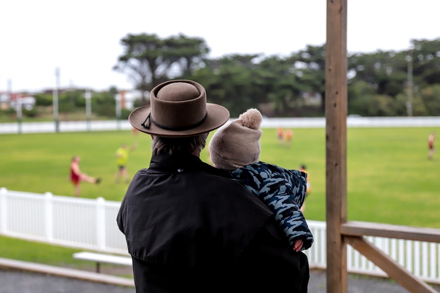 View from behind an older man wearing a wide brim hat holding a small child in his arm. They're looking out to a football ground