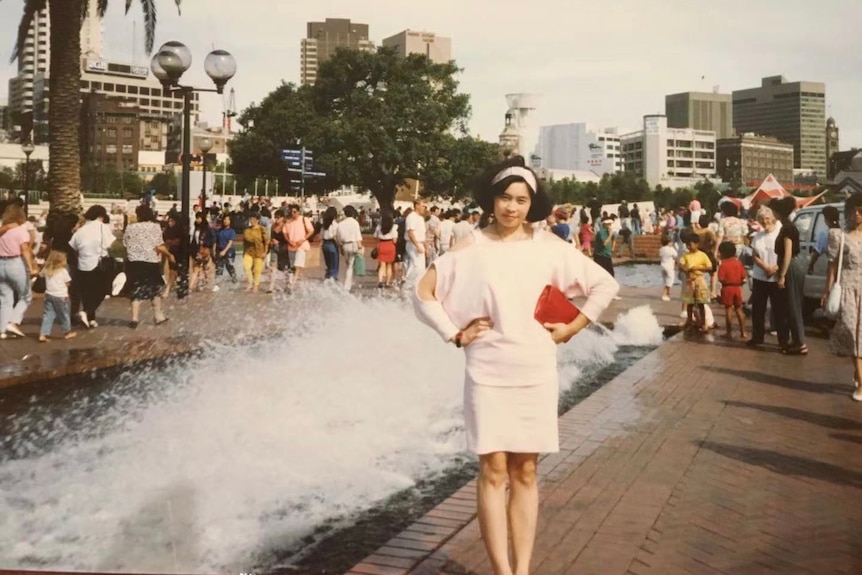 A woman in white dress standing in front of a fountain holding a red purse.