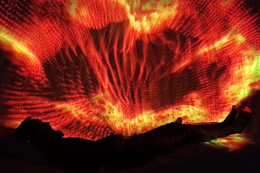 Participant lies in interactive bed wearing VR headset, surrounded by kaleidoscopic visuals.