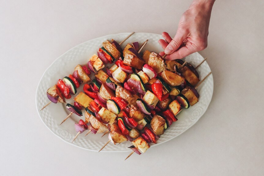 A white oval dish filled with baked vegetable and haloumi skewers. A hand reaches for one of the skewers.