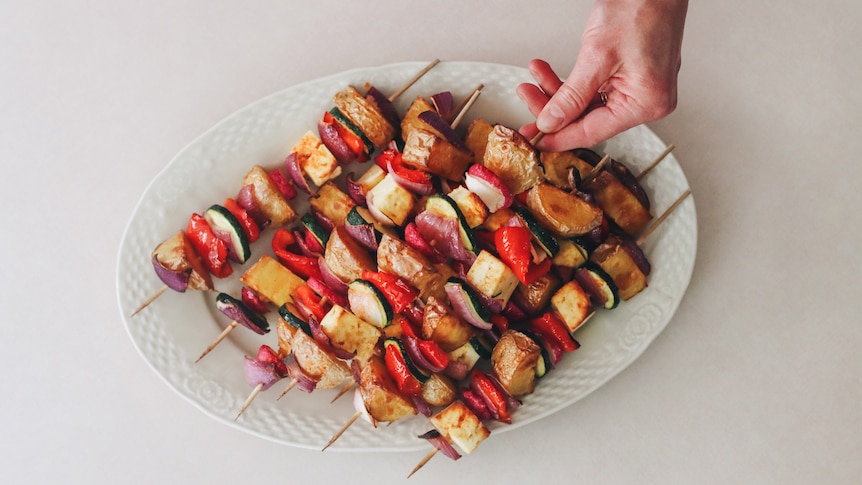 A white oval dish filled with baked vegetable and haloumi skewers. A hand reaches for one of the skewers.