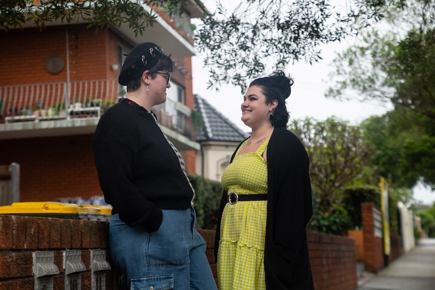 Two people stand and smile at each other outside a red brick apartment block.