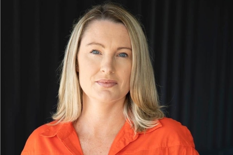An attractive blonde woman in an orange shirt looks into the camera in front of a black background