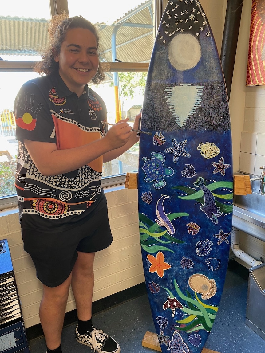 Slater Simpson wearing a shirt printed with the Aboriginal flag and art, smiling and standing next to a painted surfboard.
