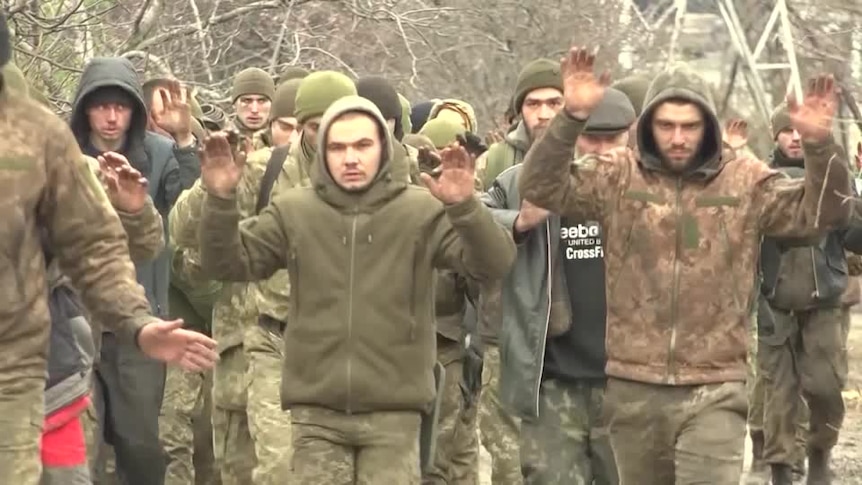 A group of men in camaflouge walk with their hands in the air