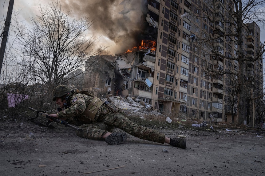 Ukrainian soldier takes cover on ground in front of burning building hit by air strike.