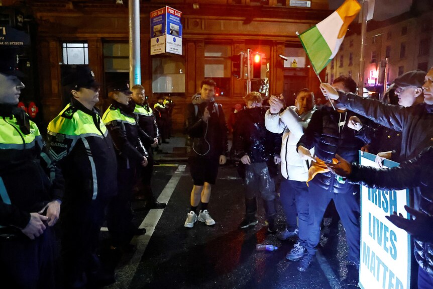 Police with riot shields and protestors holding an Irish flag stand across from each other.