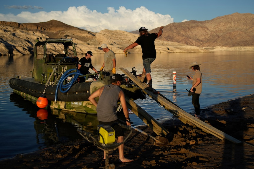 A man walks up a board to a boat on water edge. Two more men are on boat and two are on shoreline. Mountains and sky behind.