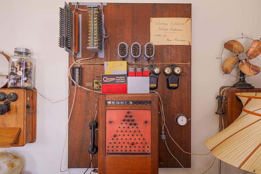 An old antique telephone hangs on a wooden board next to a large vintage lampshade and wooden fan.