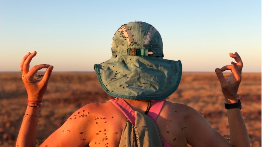 A woman faces away from the camera, holding her arms up to show dozens of flies stuck to her skin