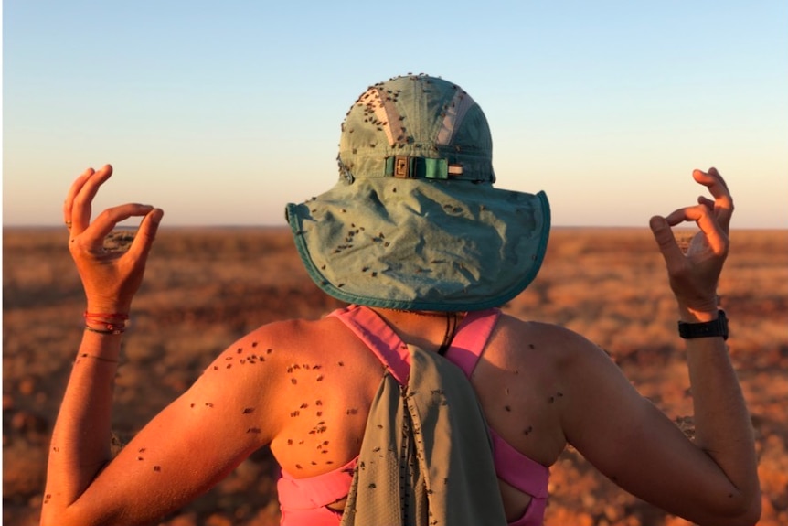 A woman faces away from the camera, holding her arms up to show dozens of flies stuck to her skin