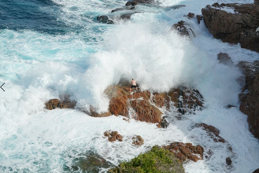 A man standing on a rock with a wave crashing over top of it