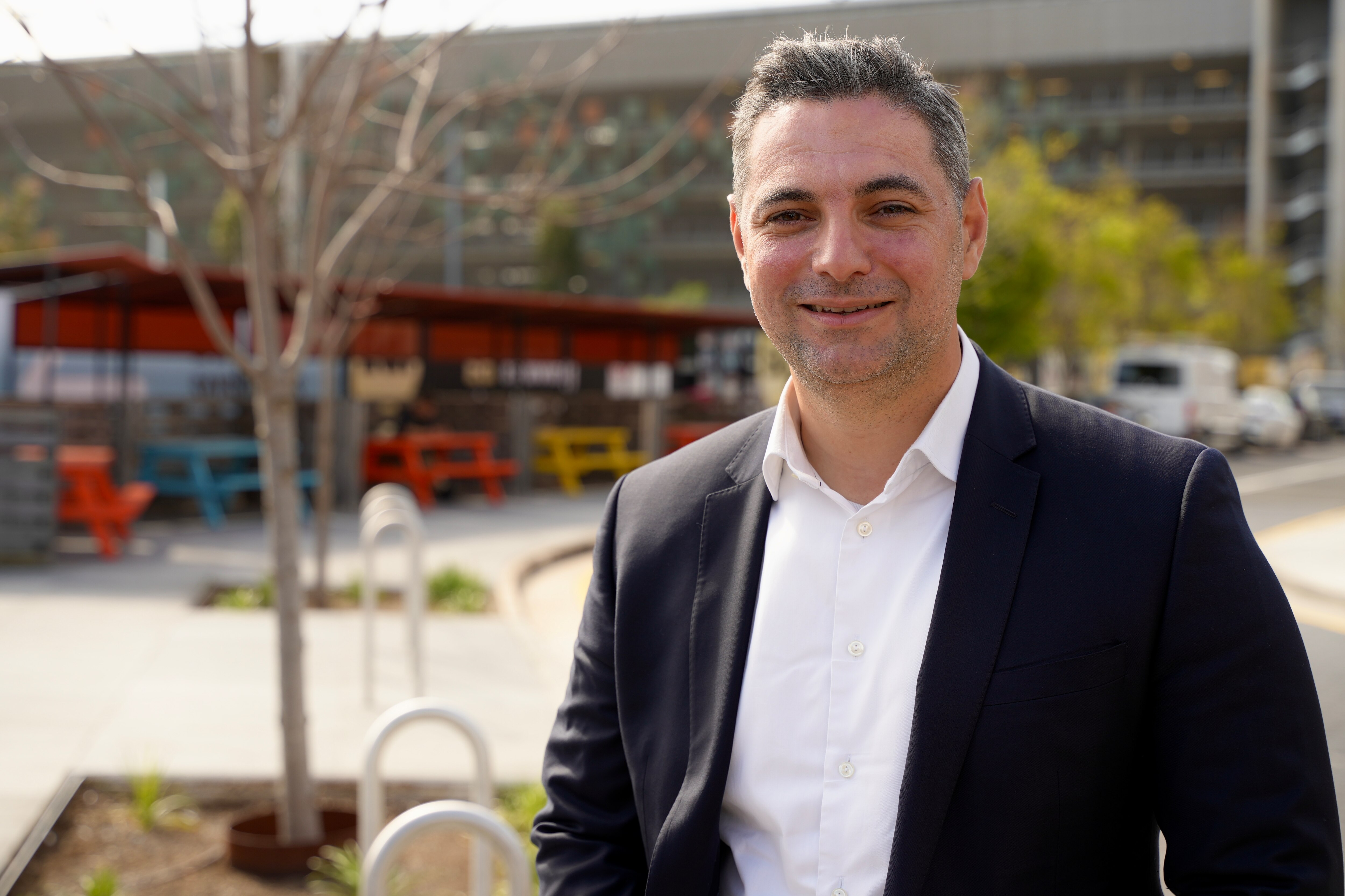Standing on the side of a road. Medium closeup framed to the right with trees and coloured park benches in the distance.