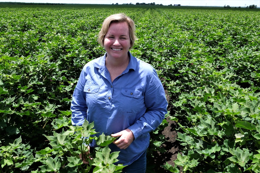 A woman with short blonde hair grins in a bright green field of cotton that's grown to waist height.