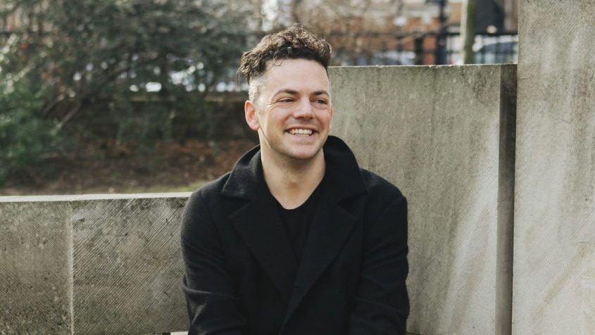 Man in black with cropped curly hair laughing while seated on concrete blocks.