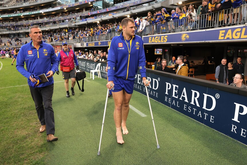 A West Coast AFL player goes round the boundary on crutches as a trainer watches.