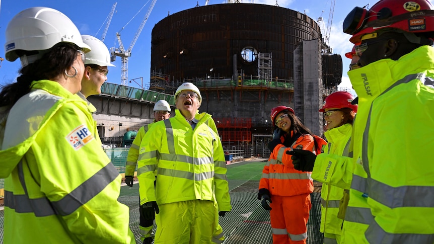 a group of people in hi vis at a nuclear power plant surround Boris Johnson who is wearing hi vis with his mouth open looking up