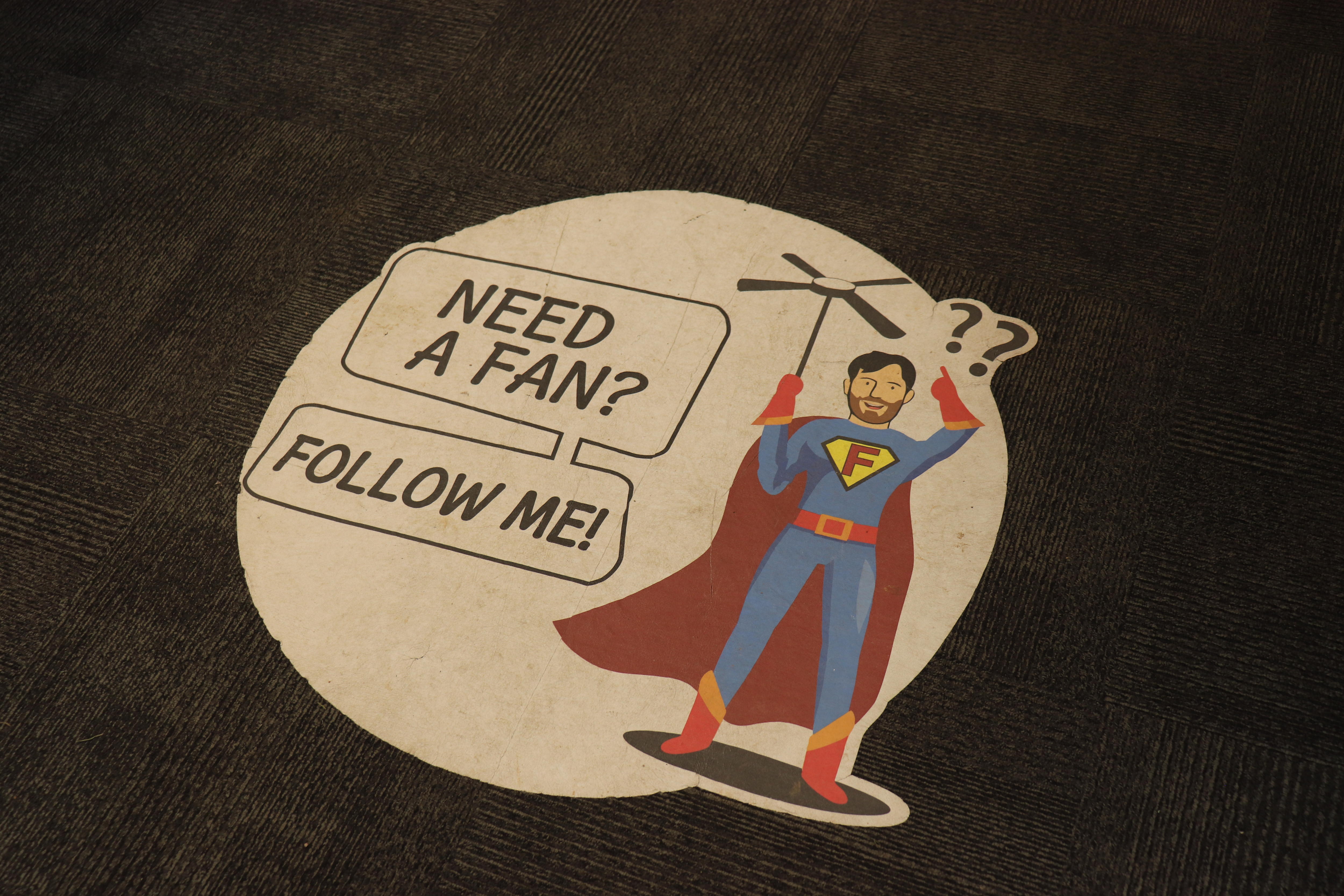 A sign on the floor says "Need a fan? Follow me" next to a superhero in a blue suit with a red cape.