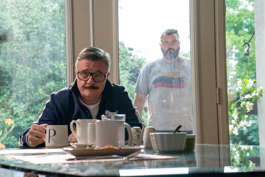 A film still of Nathan Lane and Denis Ménochet. Lane sits at a breakfast table in a tracksuit, Ménochet is behind a glass door.