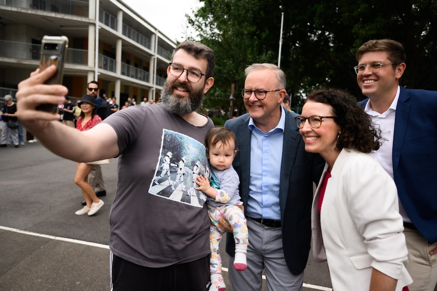 a man with a beard holding a baby taking a selfie with a group of people