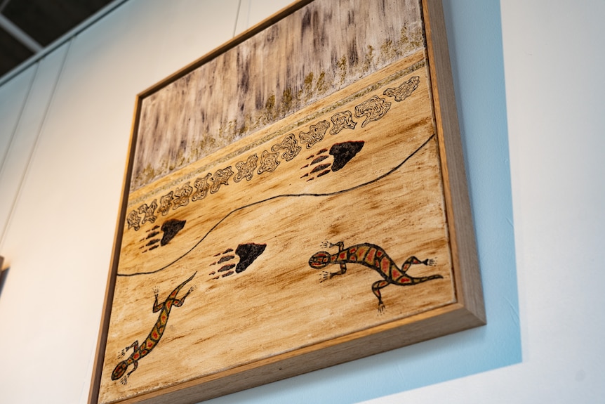 A painting with earthen colours shows animal footprints and two lizard-like animals hangs on a white wall.