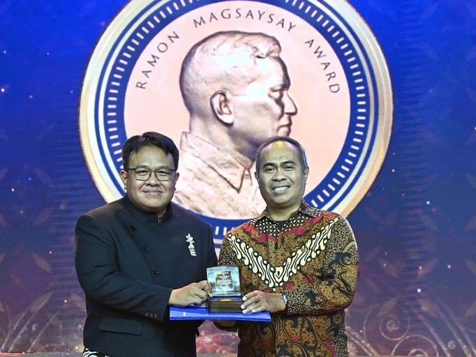 Two men holding a trophy looking at the camera, the one on the left is wearing black, the other is wearing batik.