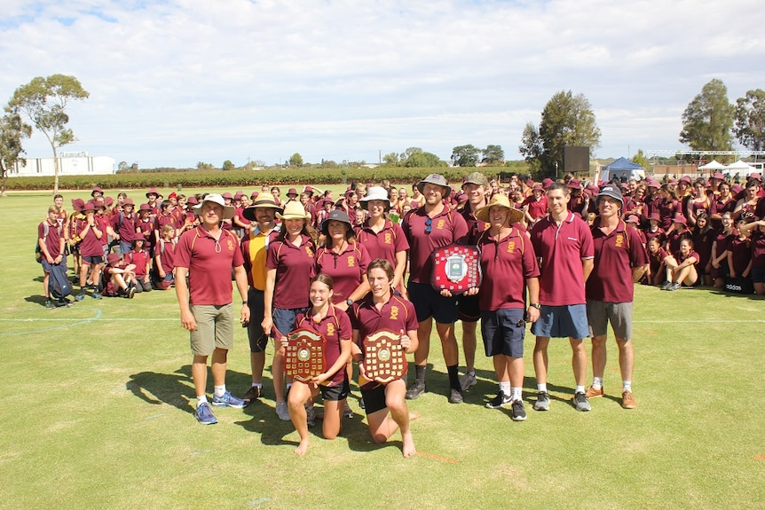 A large group of people dressed in maroon are standing on a school oval. The people at the front are holding award shields.