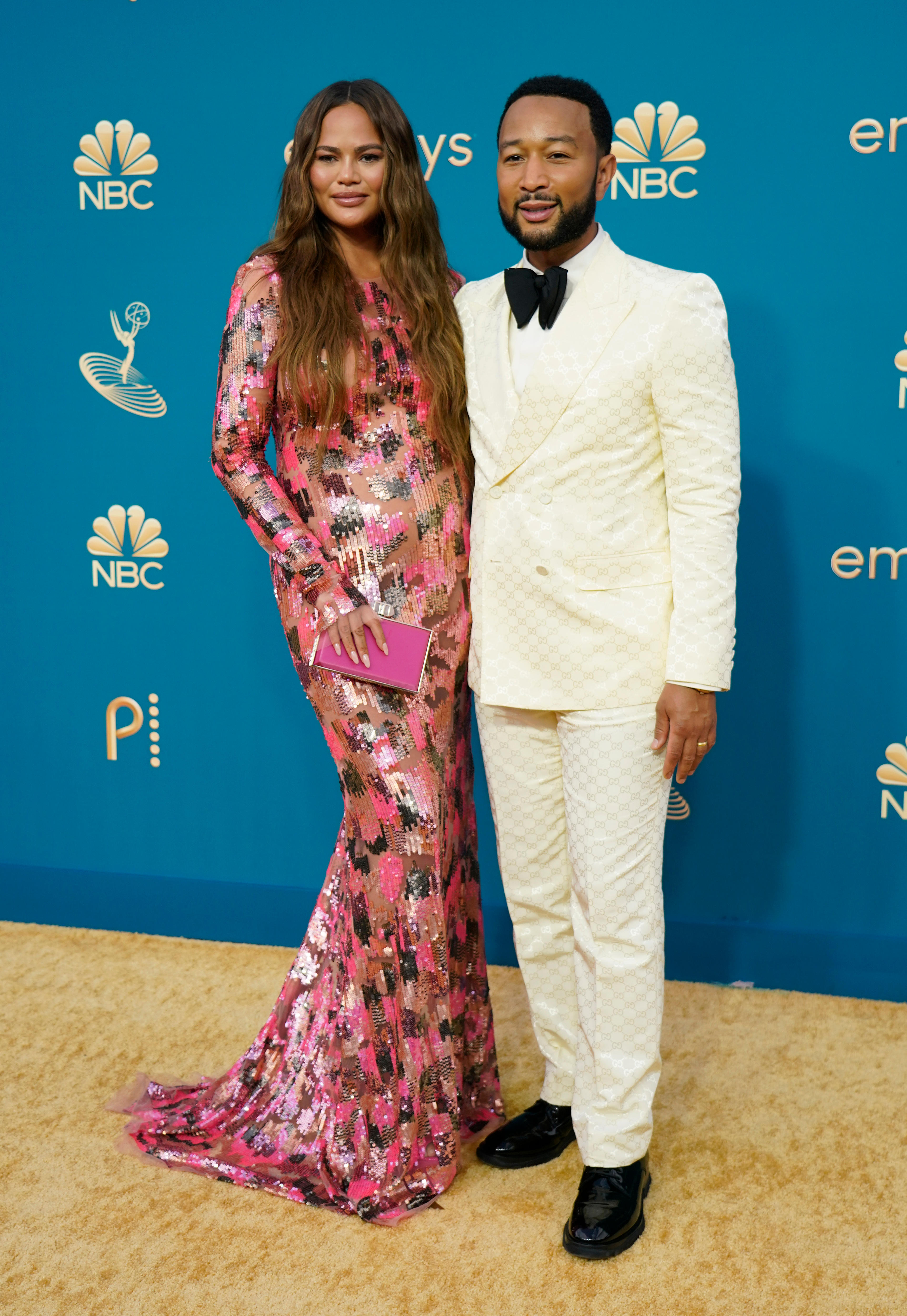 Chrissy Teigen wears a long sleeved pink patterned gown and John Legend wears a white suit with a black bow tie
