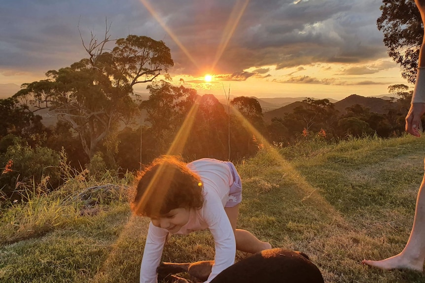 Con la puesta de sol de fondo, una joven es levantada en el aire por las manos de un adulto.