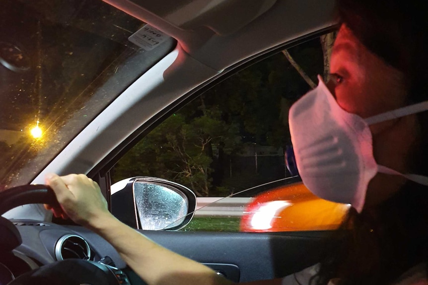 A woman drives a car at night while wearing a facemask.