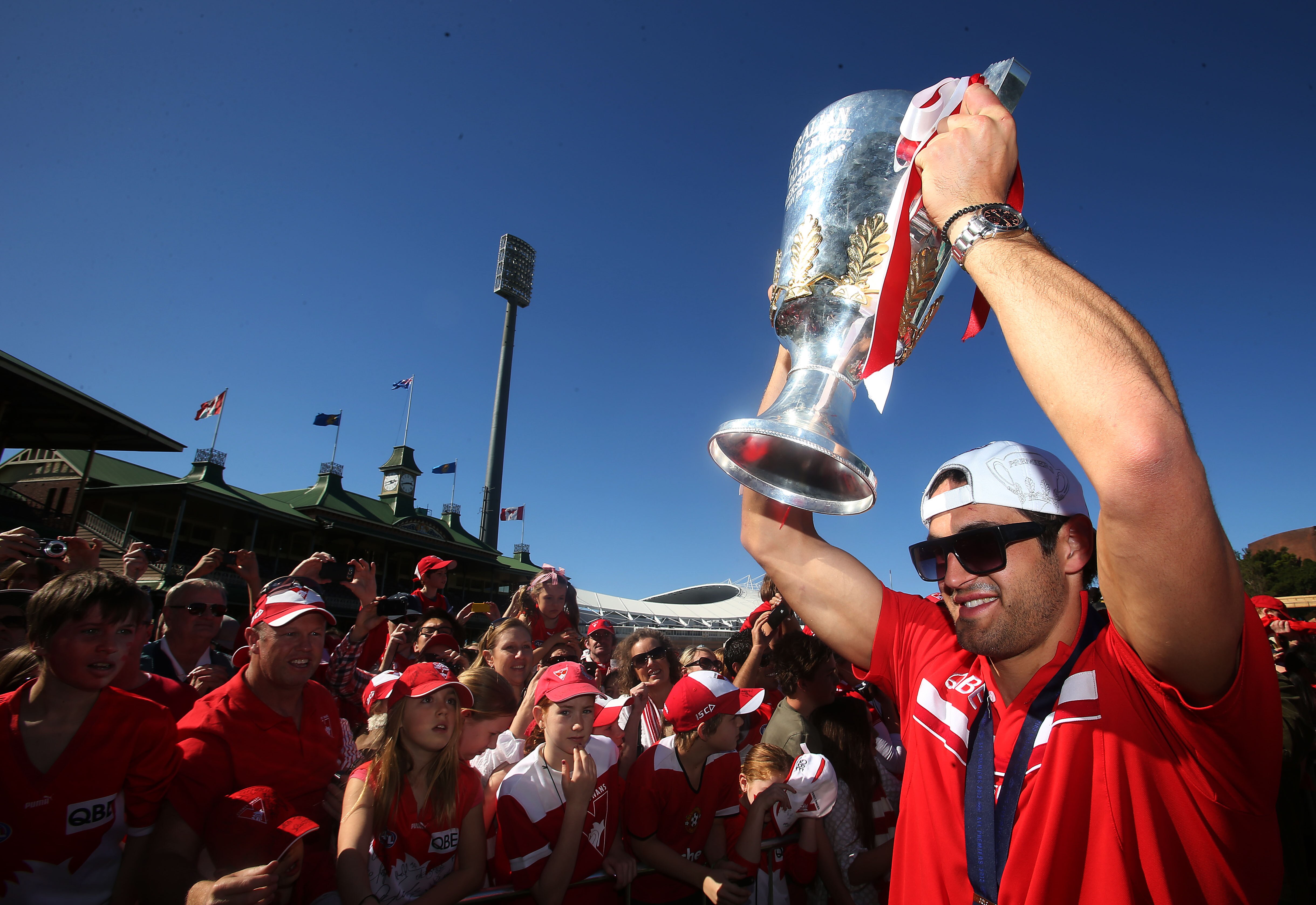 Josh Kennedy holds the premiership cup above his head in front of Swans fans, while wearing sunglasses and a backwards cap