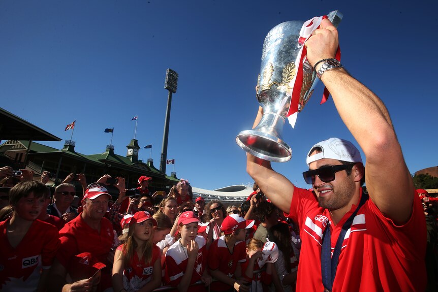 Josh Kennedy holds the premiership cup above his head in front of Swans fans, while wearing sunglasses and a backwards cap