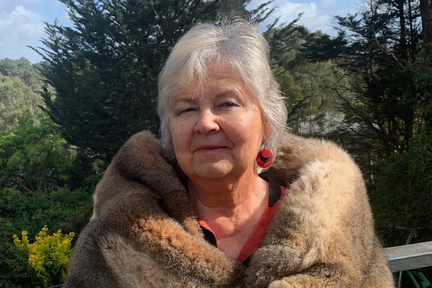 Loraine Padgham stands wrapped in a possum-skin cloak outdoors, in front of rolling green hills and bush.