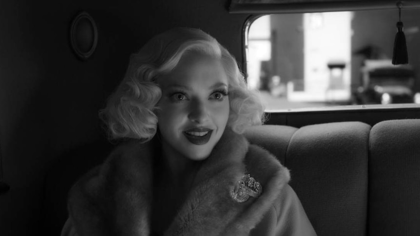 Black and white close-up of actress in platinum blonde curly wig and wearing coat with fur collar sitting in the back of a car.