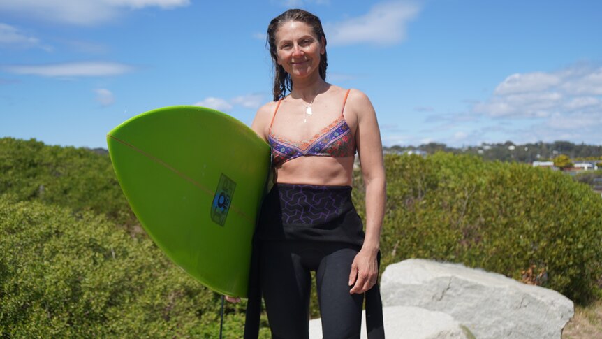 A woman smiles at a camera half in a wetsuit and bikini with a green surfboard.
